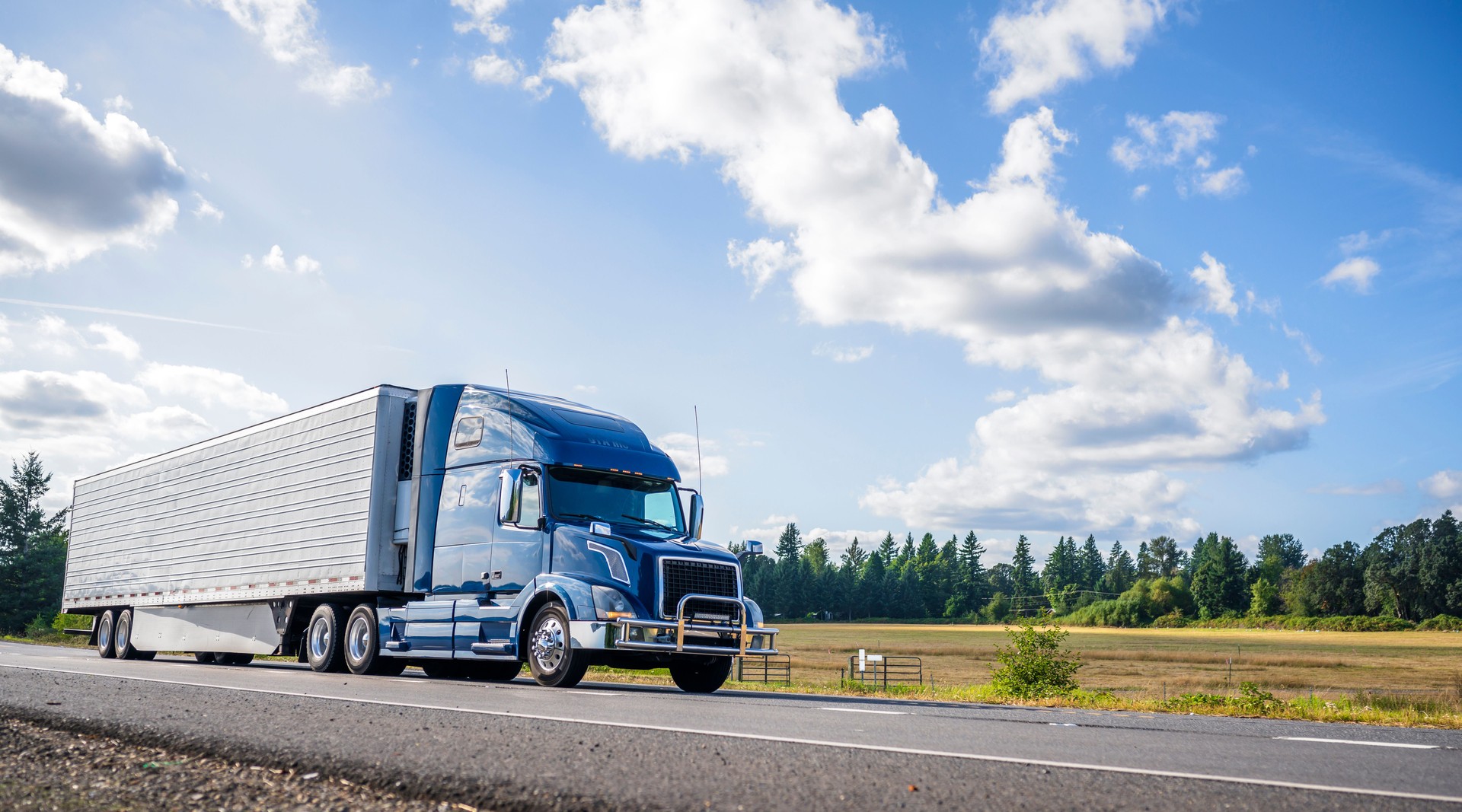 Blue big rig semi truck with grille guard trabsporting frozen cargo in refrigerator semi trailer with skirt spoiler running on the straight road